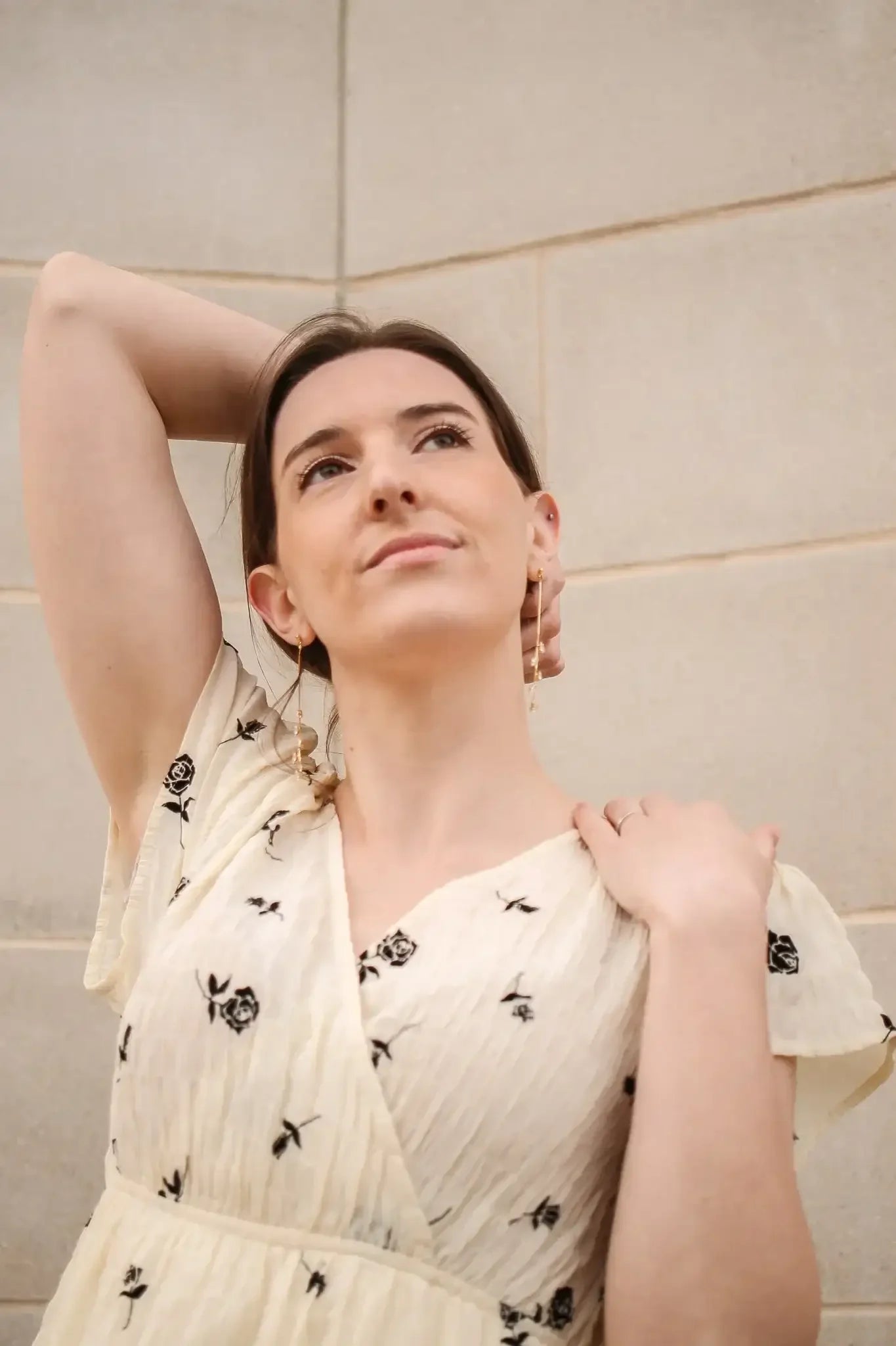 A woman in a floral dress raises one arm, casually resting her hand on her neck while gazing slightly upwards, set against a smooth stone wall background.