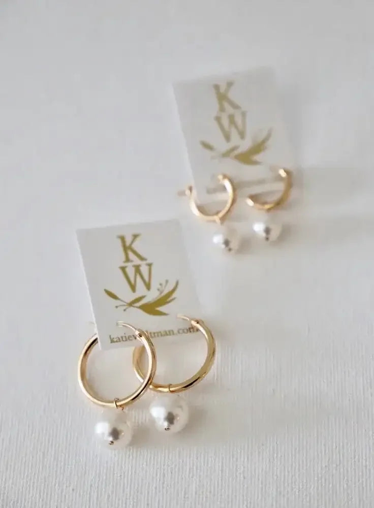 Gold hoop earrings with white pearls are displayed on a white background alongside a branded card featuring "KW" and the website "katiebutman.com." The earrings hang beneath the card.