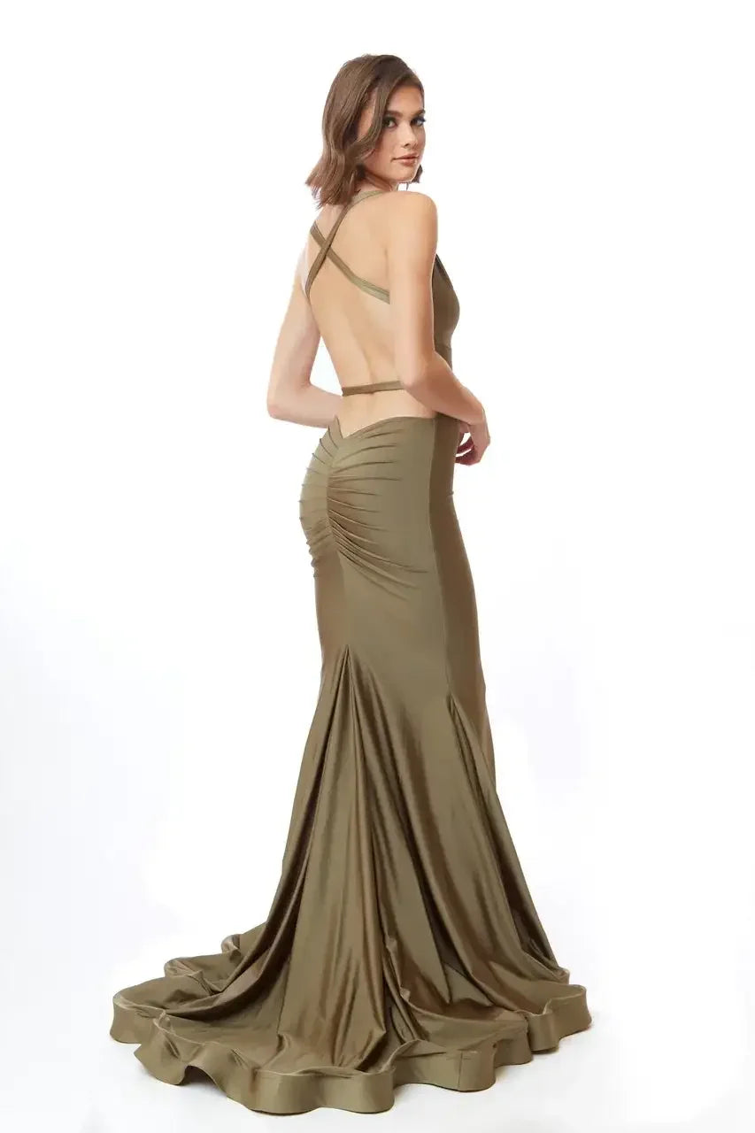A woman in a form-fitting, olive-green gown stands elegantly, showcasing a low back and crossed straps. The dress flows beautifully, trailing slightly on the floor, set against a white background.