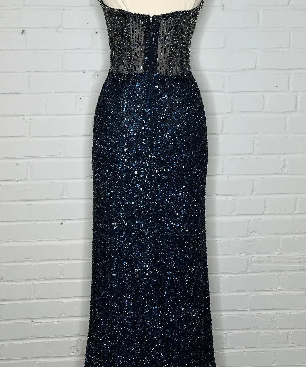 A shimmering, floor-length navy gown adorned with sequins and a transparent bodice is displayed on a dress form against a white brick wall background. The dress exudes elegance and glamour.