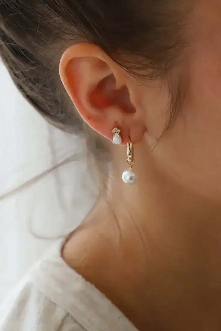 A close-up of a person's ear adorned with two earrings: one a small opal stud, the other a gold hoop holding a pearl, set against a soft, light backdrop.