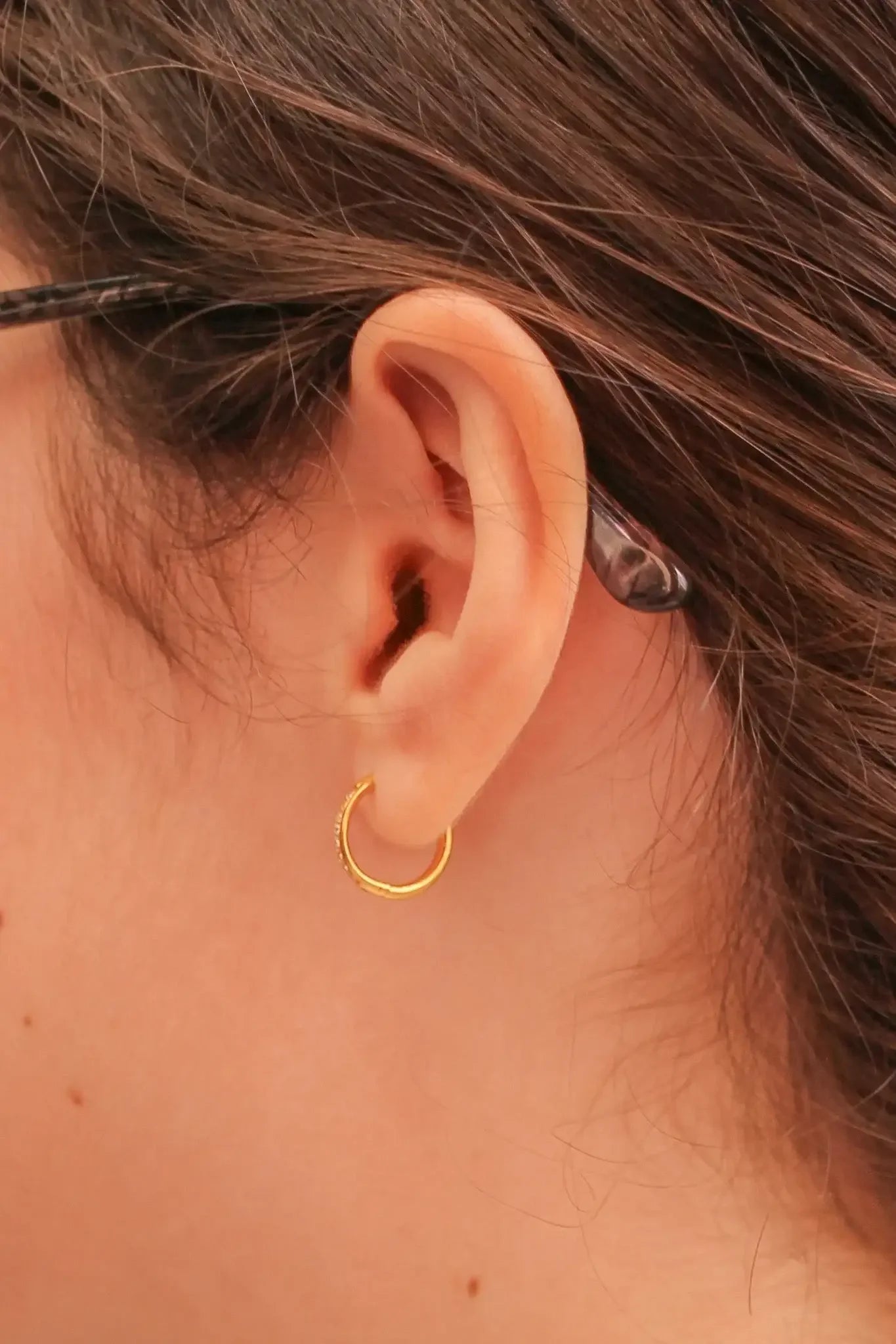A close-up of a human ear adorned with a small gold hoop earring, surrounded by brown hair, creating an intimate view of jewelry in a personal context.