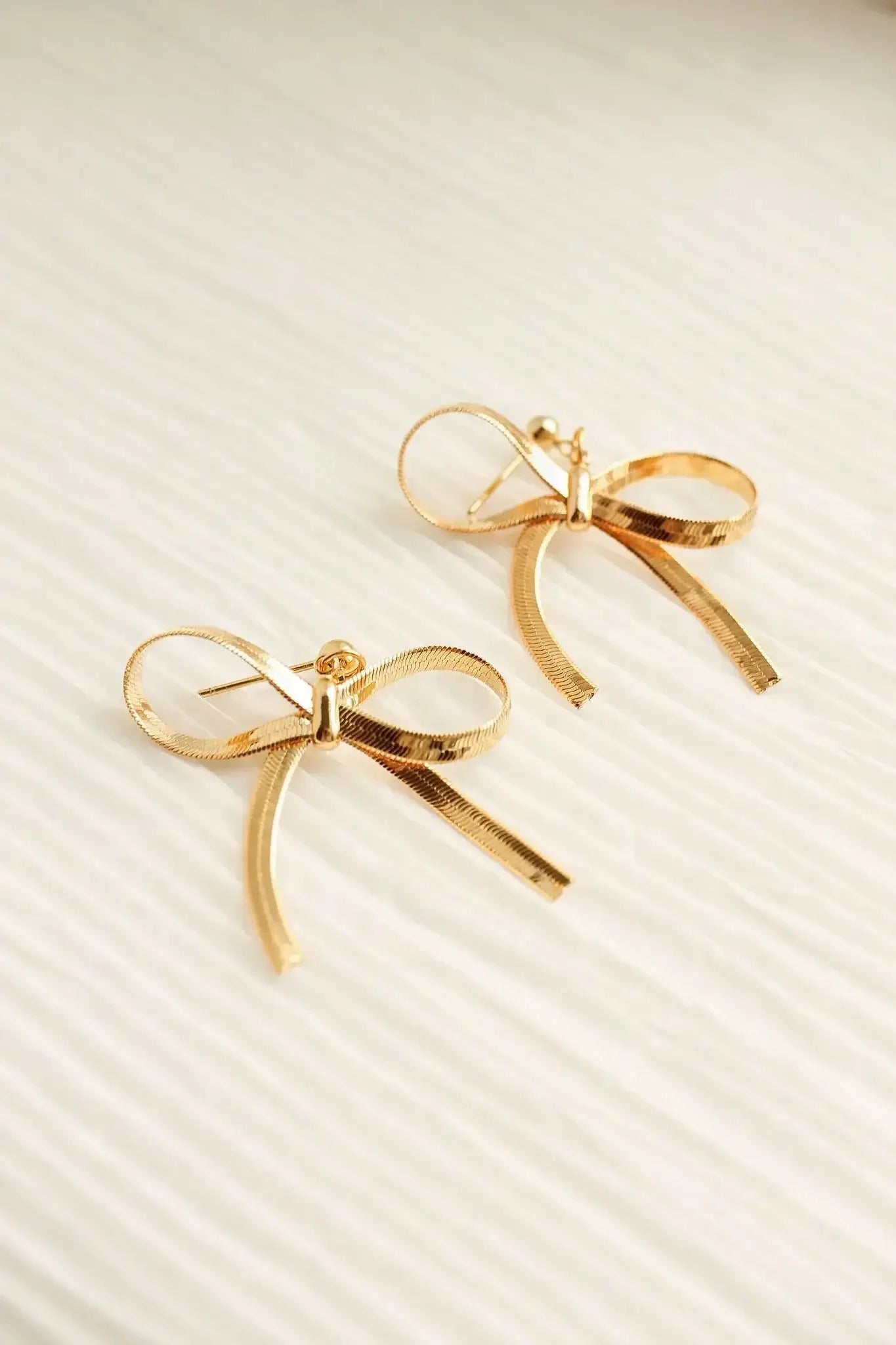 Two golden bow-shaped earrings rest on a soft, textured surface. Their design features loops and tails, reflecting light elegantly, suggesting a delicate and stylish accessory.