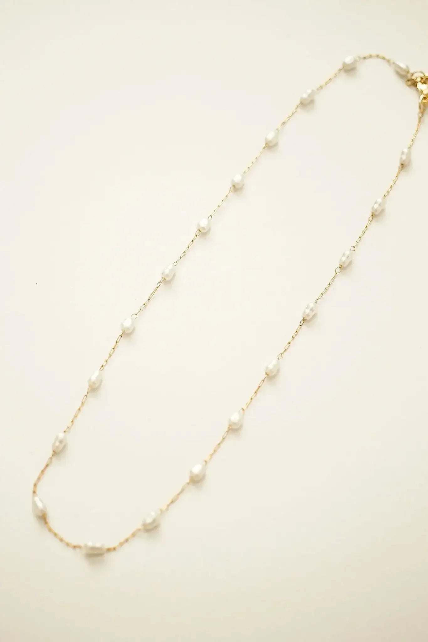 A delicate gold chain necklace features evenly spaced small white pearls. It is laid flat against a soft, light-colored background, emphasizing its elegance and simplicity.