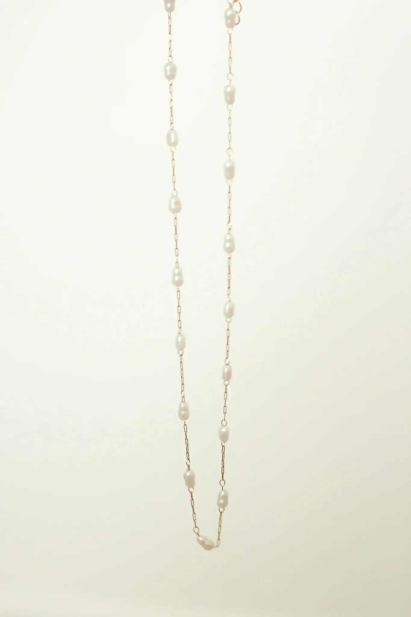 A delicate pearl necklace hangs vertically, with evenly spaced pearls linked by fine gold chain, against a soft, light-colored background, highlighting its elegance and simplicity.