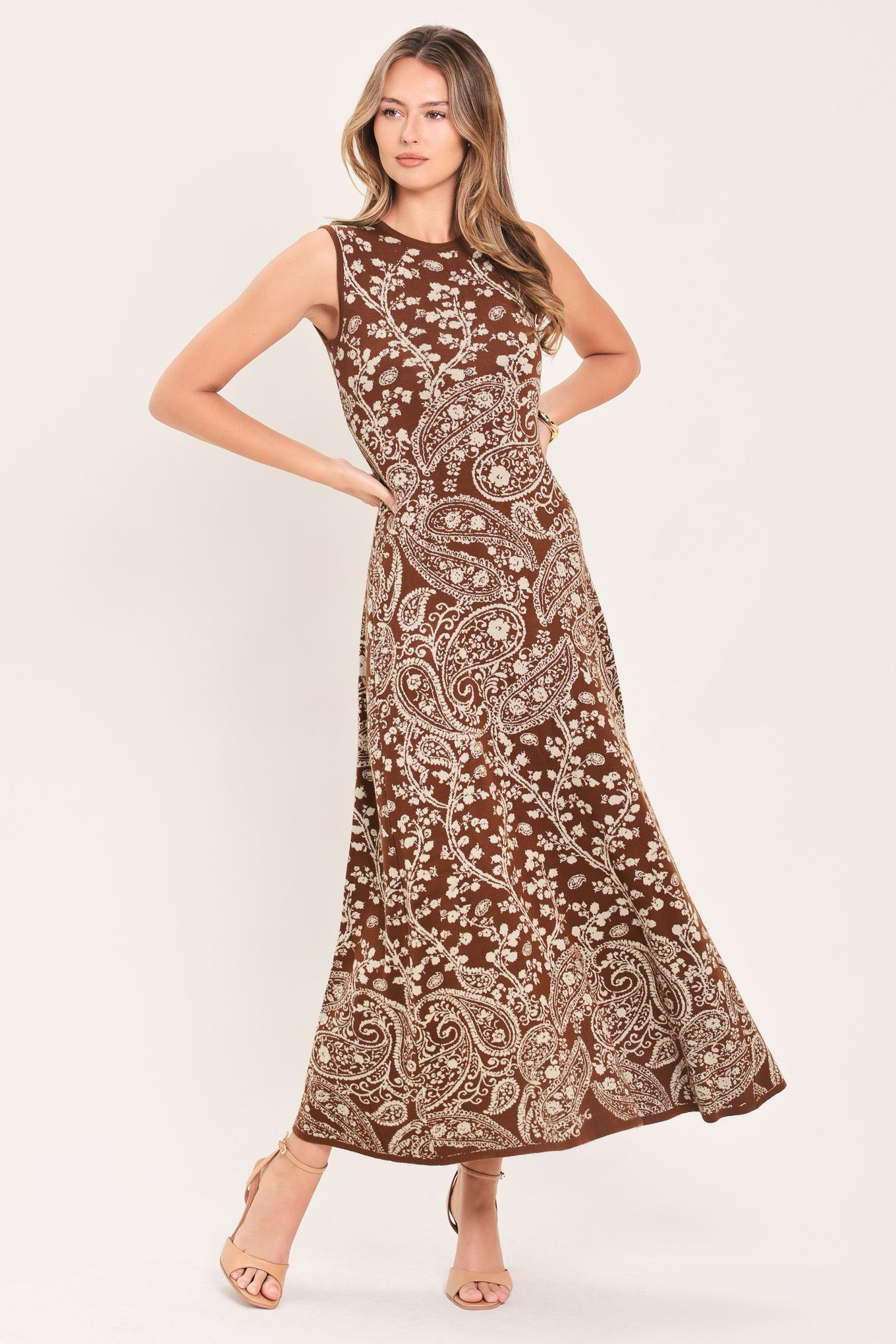 HAZEL PAISLEY EMBRACE JACQUARD SWEATER MIDI DRESS | Lady Black Tie | A woman modeling a sleeveless, floor-length brown maxi dress with an intricate beige paisley and floral pattern, posed against a solid off-white studio background.