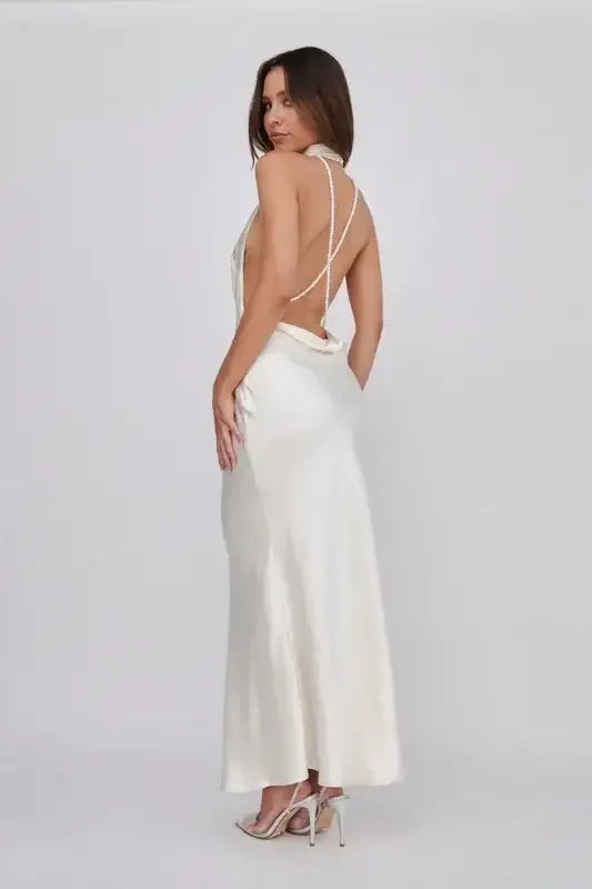 A woman stands elegantly in a white, backless dress with thin straps. She poses sideways, showcasing the dress's sleek design against a plain, light background, enhancing its sophistication.