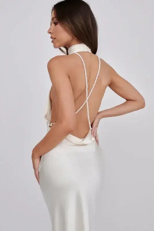 A woman poses elegantly in a sleek, backless white dress adorned with pearl straps. Her long hair cascades over her shoulder against a neutral backdrop, showcasing the dress's refined style.