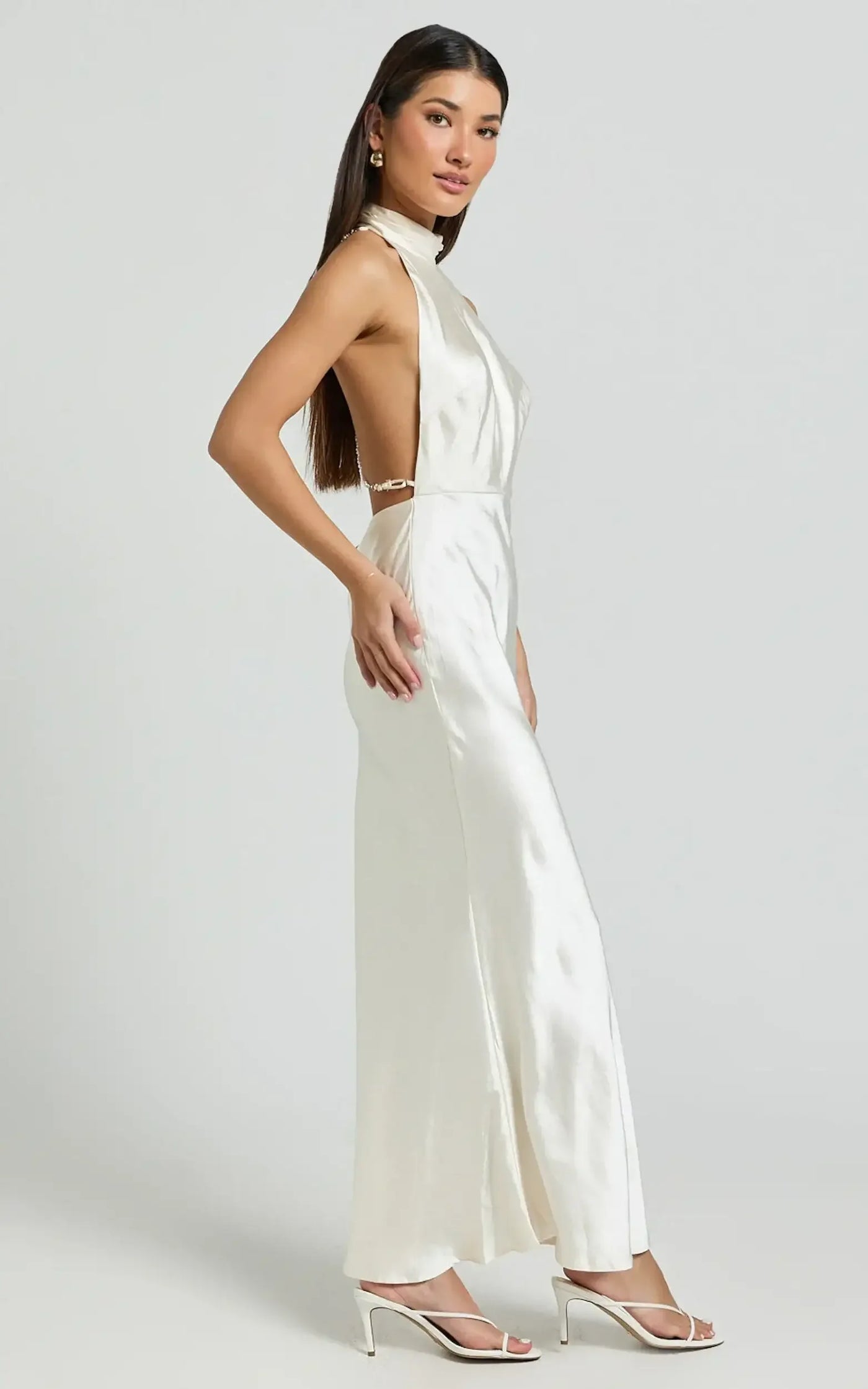 A woman wearing a sleek, white satin gown stands gracefully, showcasing a high neckline and open back. She poses in a minimalistic, neutral background, complemented by stylish heels.