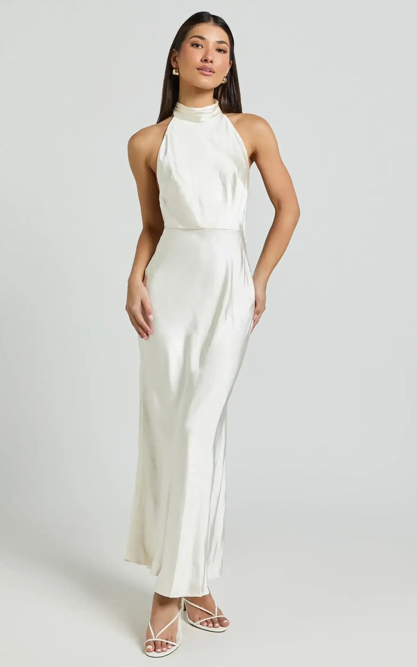 A woman stands elegantly in a long, white halter dress, showcasing confident posture against a light gray background. Her stylish strappy sandals complement the outfit, enhancing her sophisticated look.
