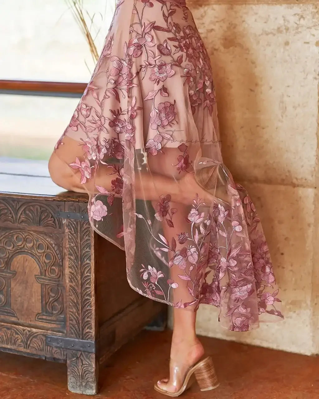 A sheer, embroidered dress is elegantly worn by a person as they gracefully lean on a wooden chest, surrounded by a softly lit interior with stone walls.