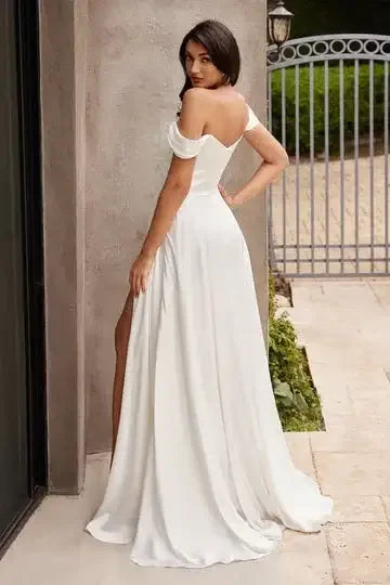 A woman in a flowing white gown stands confidently, showcasing a high slit. She poses near a stylish gate, surrounded by greenery and stone pavement.