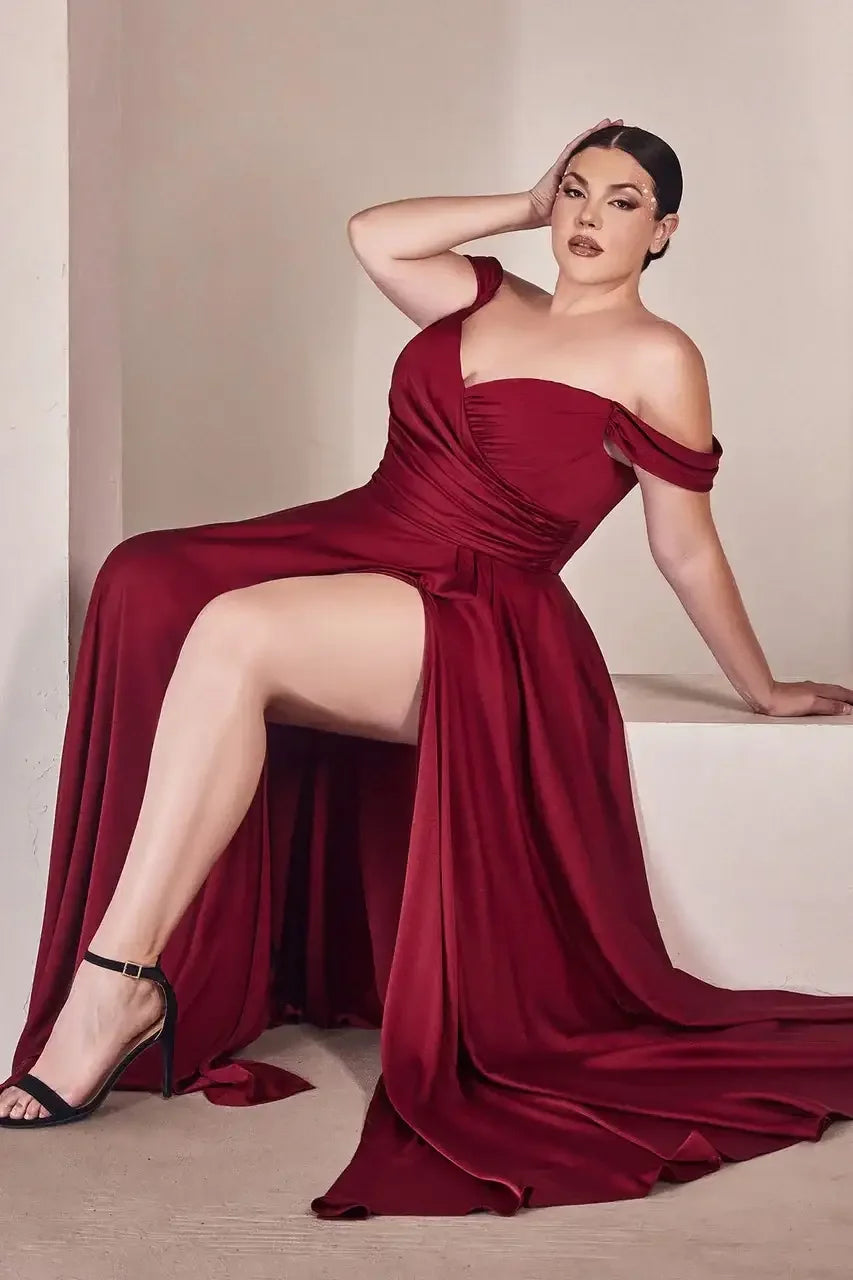 A woman in a flowing burgundy gown sits on a ledge, one hand on her head, exuding elegance and confidence in a neutral, softly lit environment.