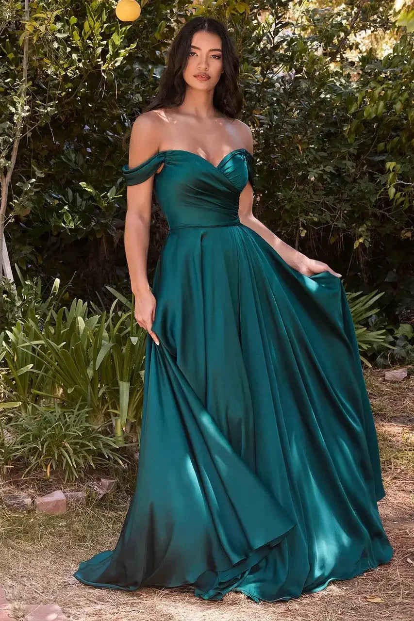 A woman stands confidently in a flowing, emerald green evening gown, holding the fabric to display its elegance, surrounded by lush greenery and natural scenery.