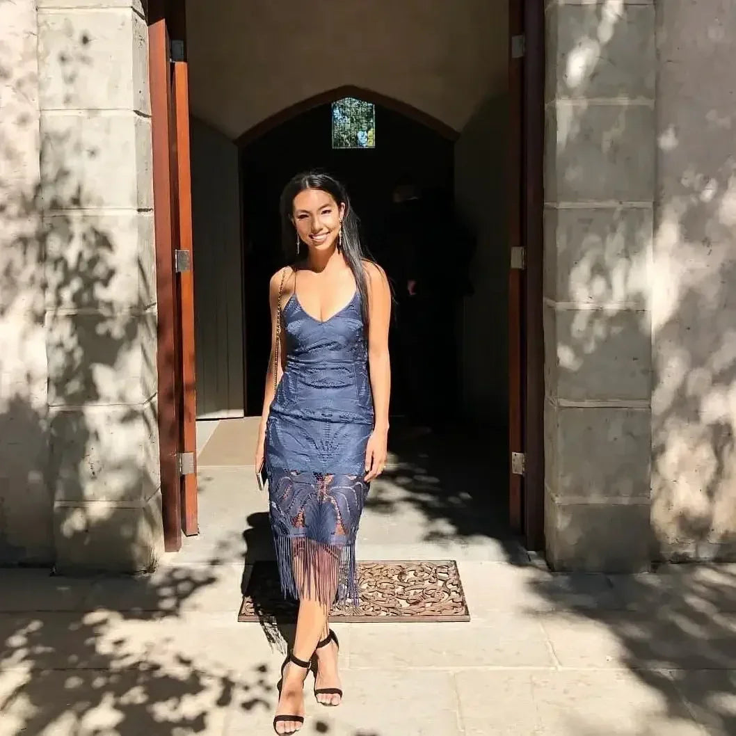A woman stands at an entrance, smiling while wearing a navy blue dress with fringe. Sunlight filters through trees, casting shadows on the stone floor, enhancing the scene's warmth.