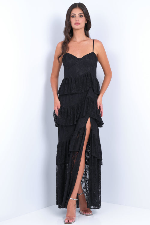 A woman stands confidently in a layered black lace dress with a thigh-high slit, on a glossy white surface, against a soft, light-colored backdrop. She wears strappy sandals.
