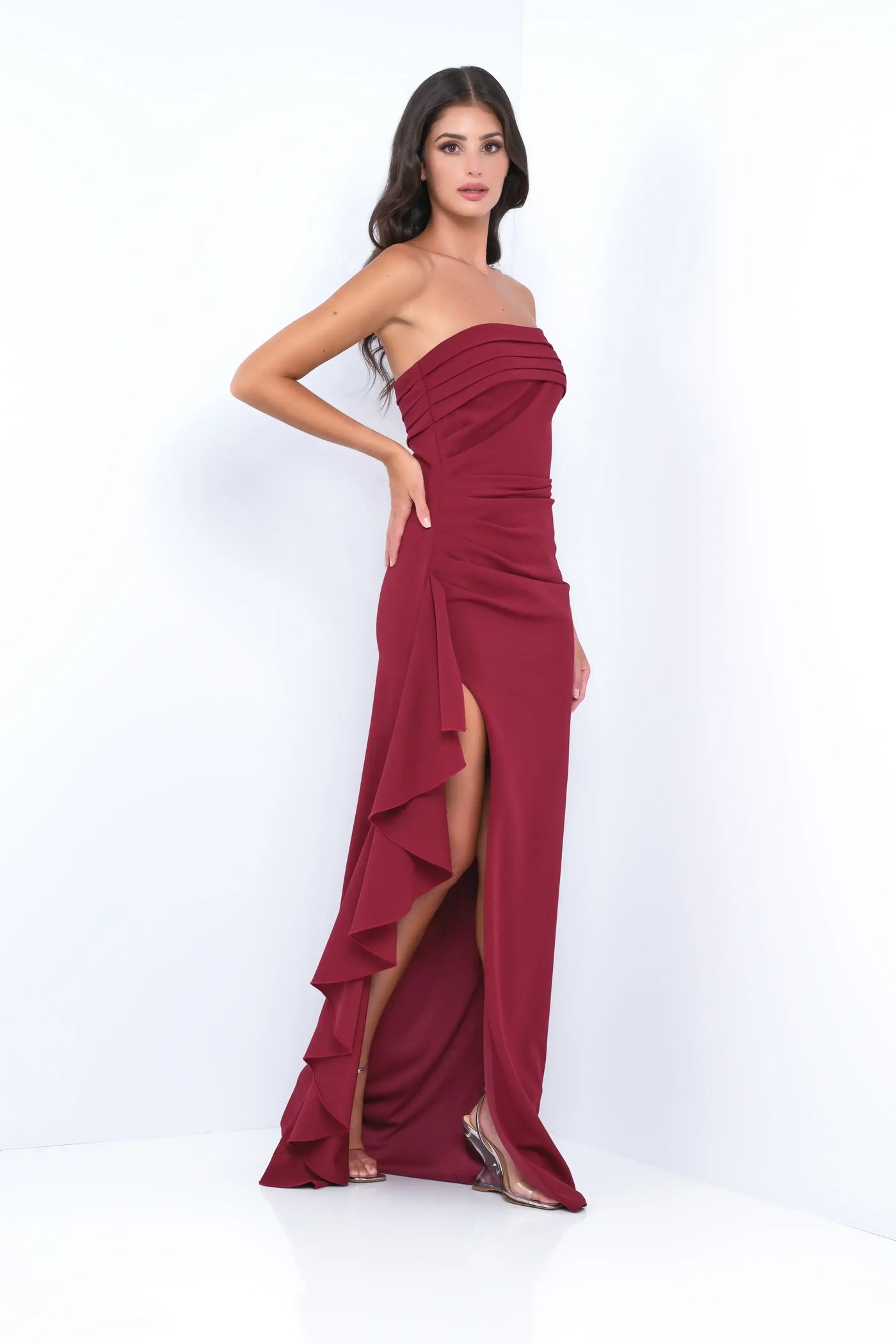 A person stands confidently in a burgundy strapless dress with pleats and a high slit, showcasing elegant ruffles. The background is minimalistic and white, enhancing the outfit's prominence.