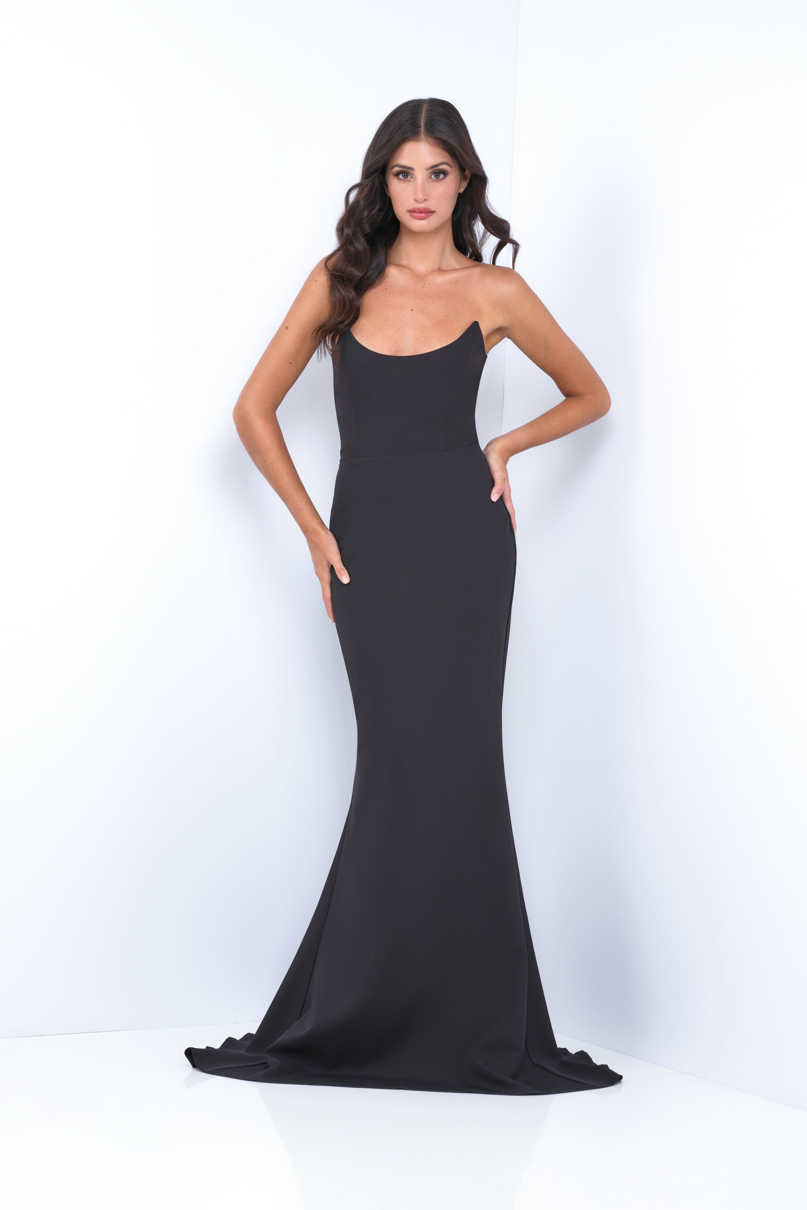 A woman is standing, wearing a sleek black evening gown that hugs her figure and flares at the bottom. The background is a simple, light-colored wall, emphasizing her elegant pose.