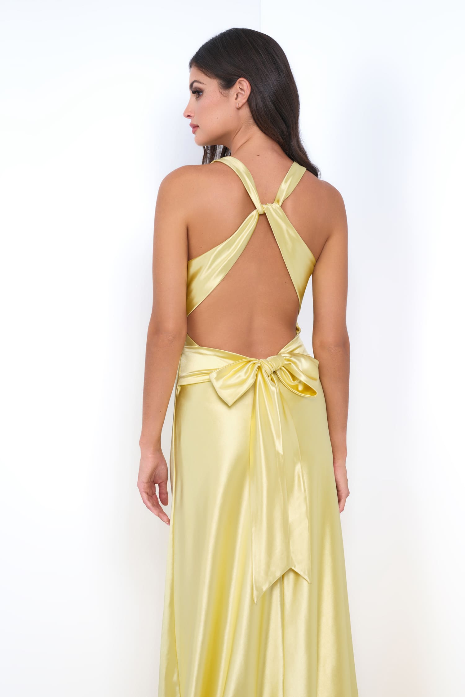 A woman modeling a yellow satin maxi dress from the back, featuring a halter-style open back with crossed straps and a large bow at the waist, set against a plain white background.