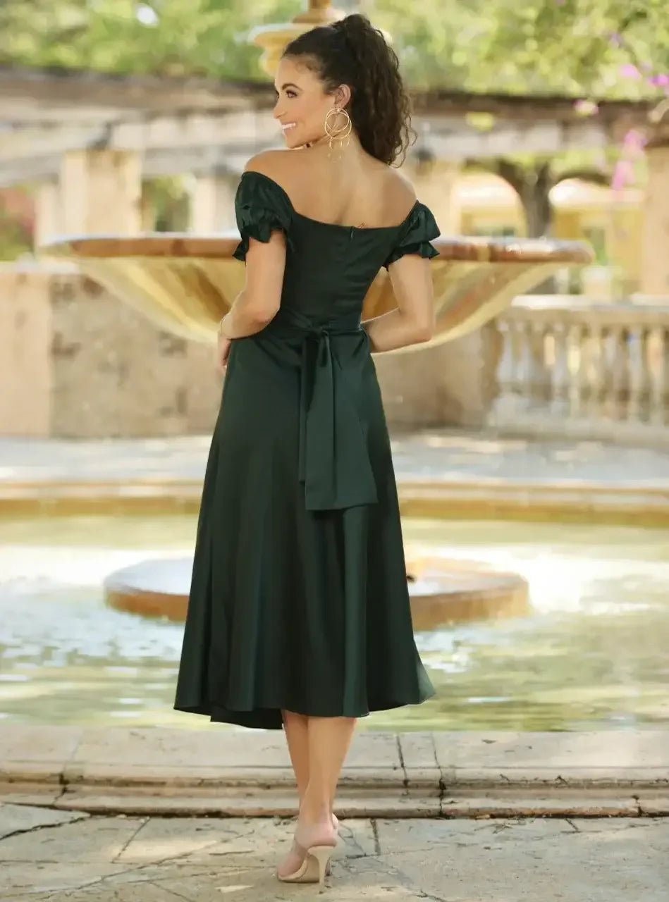 A woman in a dark green off-shoulder dress stands by a fountain, smiling and posing with her back to the camera. Sunlight filters through trees, creating a serene outdoor atmosphere.