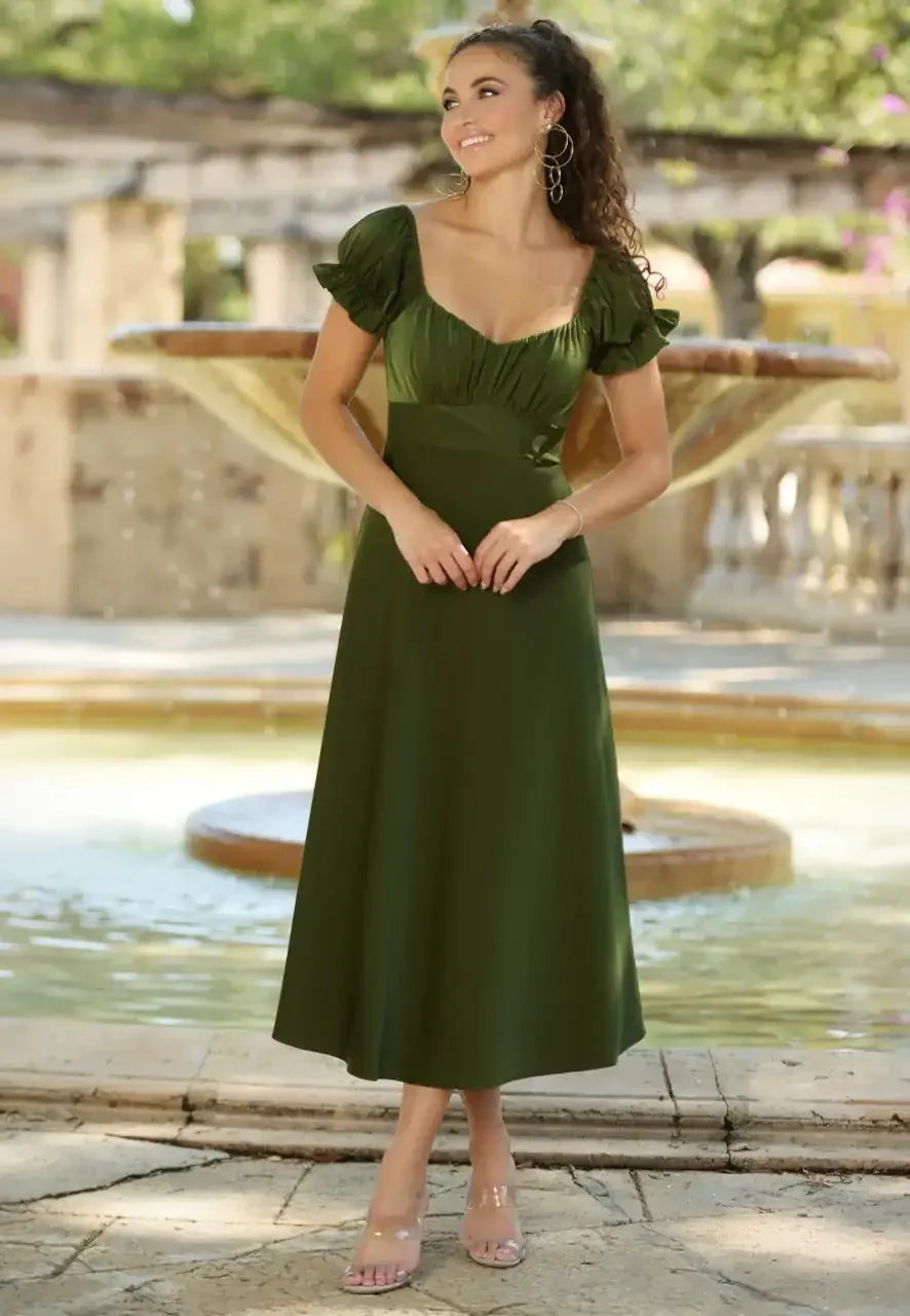 A woman stands smiling in an elegant, fitted olive green dress with puff sleeves. She is posed near a stone fountain surrounded by greenery, emulating a serene outdoor atmosphere.