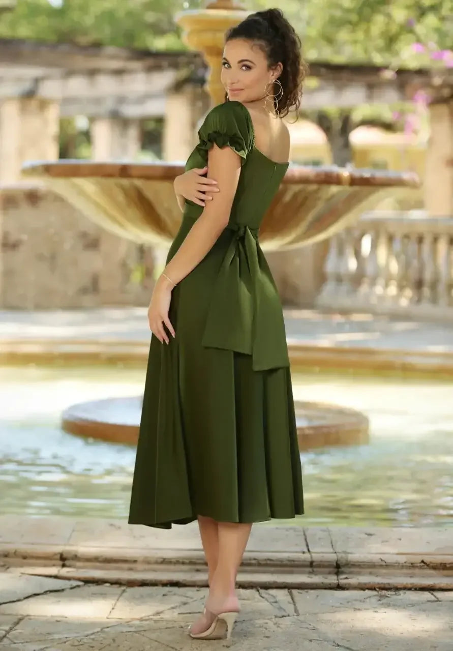 A woman in a chic, olive-green dress poses gracefully beside a fountain in a sunlit outdoor setting, smiling over her shoulder with greenery and stone architecture in the background.
