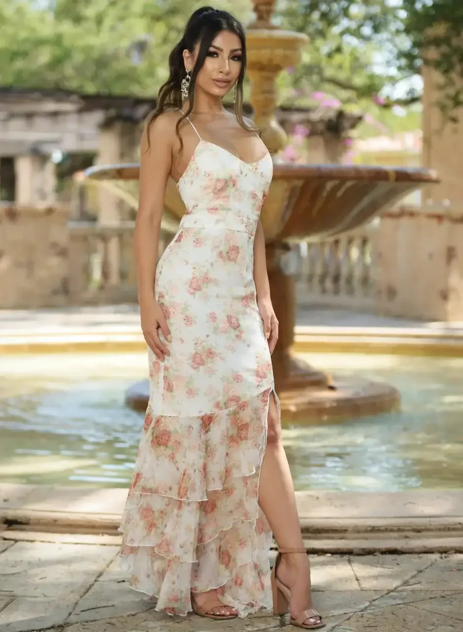 A woman in a floral dress stands beside a fountain, posing elegantly with one hand on her hip. The background features greenery and stone architecture, creating a serene outdoor setting.