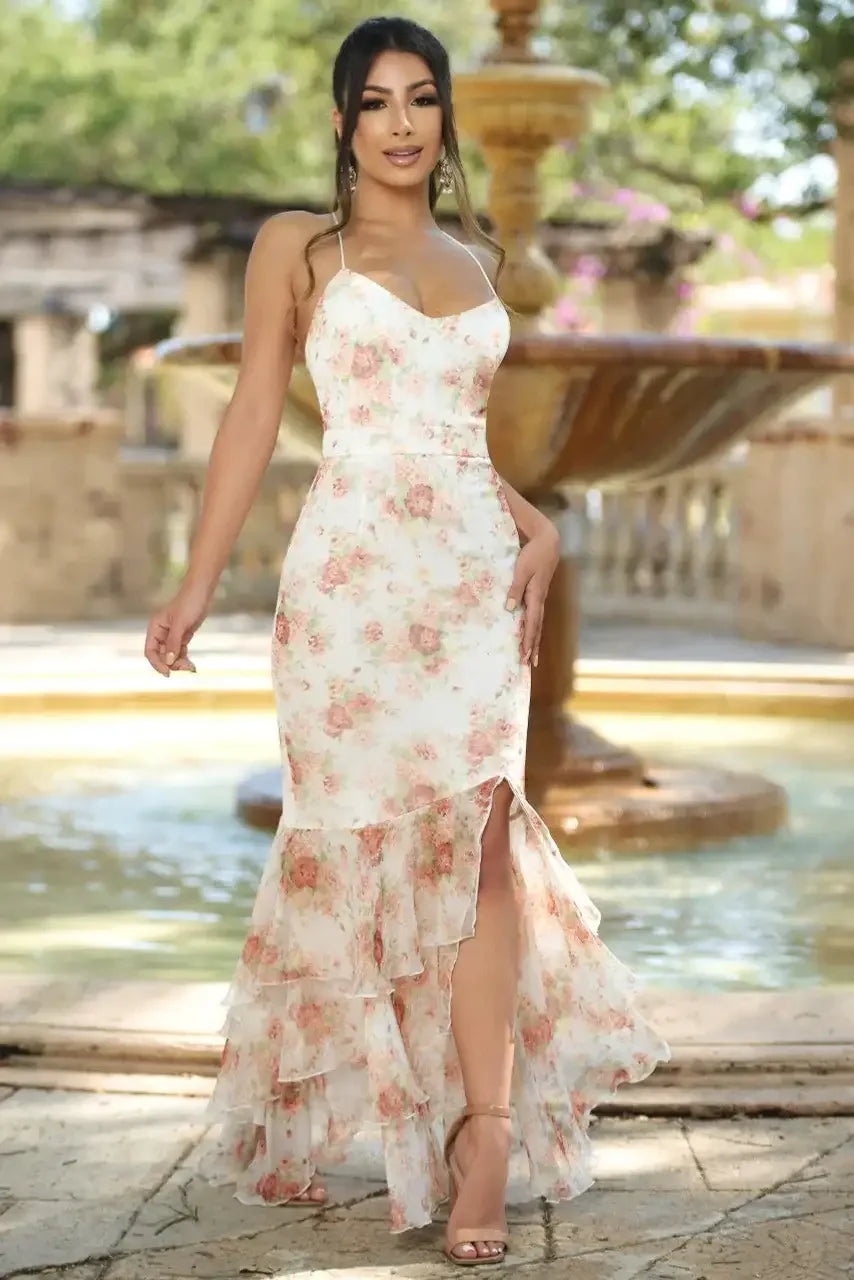 A woman stands gracefully by a fountain, wearing a floral dress with ruffles and a side slit. The sunlit garden setting is lush, enhancing her elegant pose.