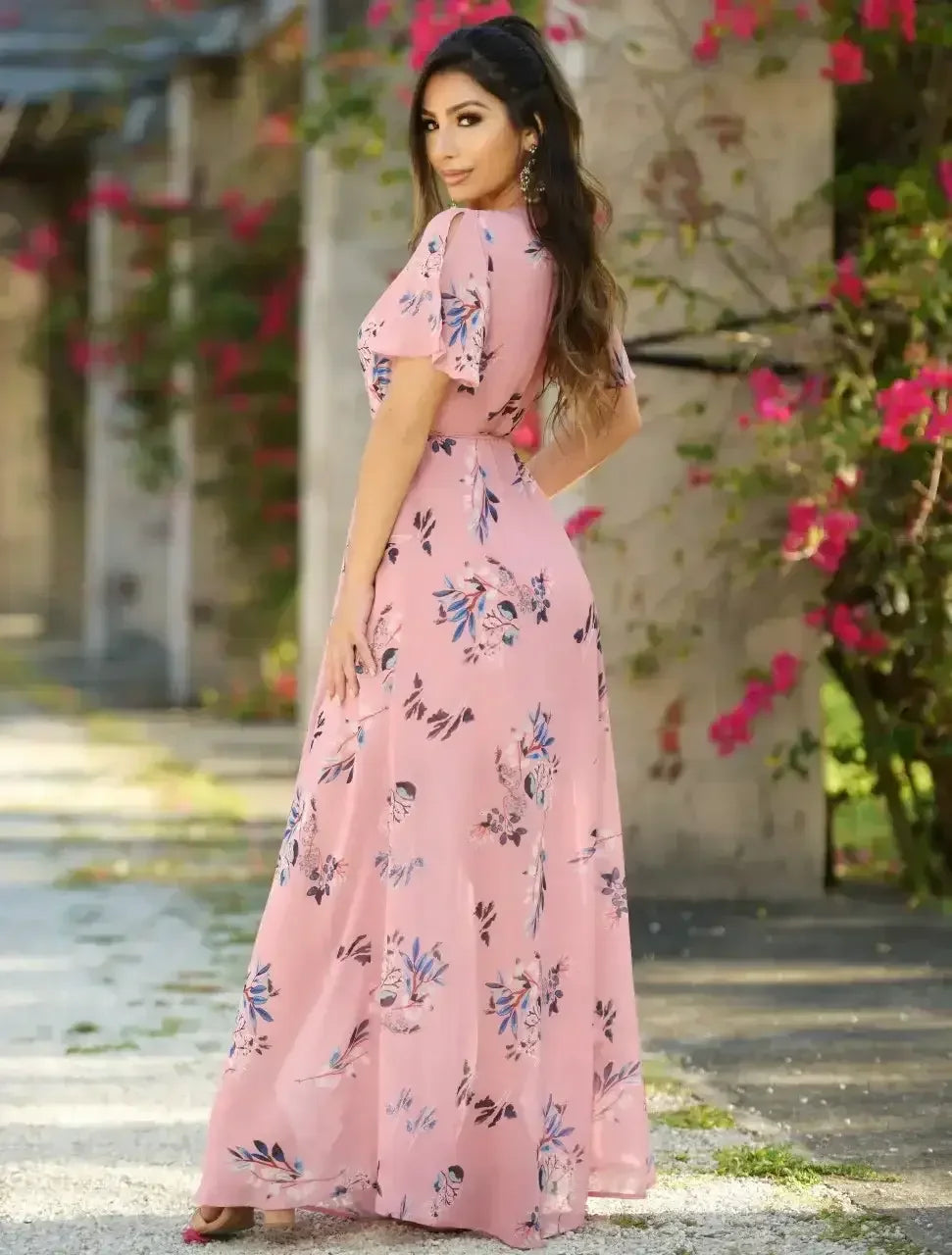 A woman in a flowing pink floral dress stands gracefully, looking over her shoulder amidst lush greenery and blooming flowers, with a stone path extending behind her.