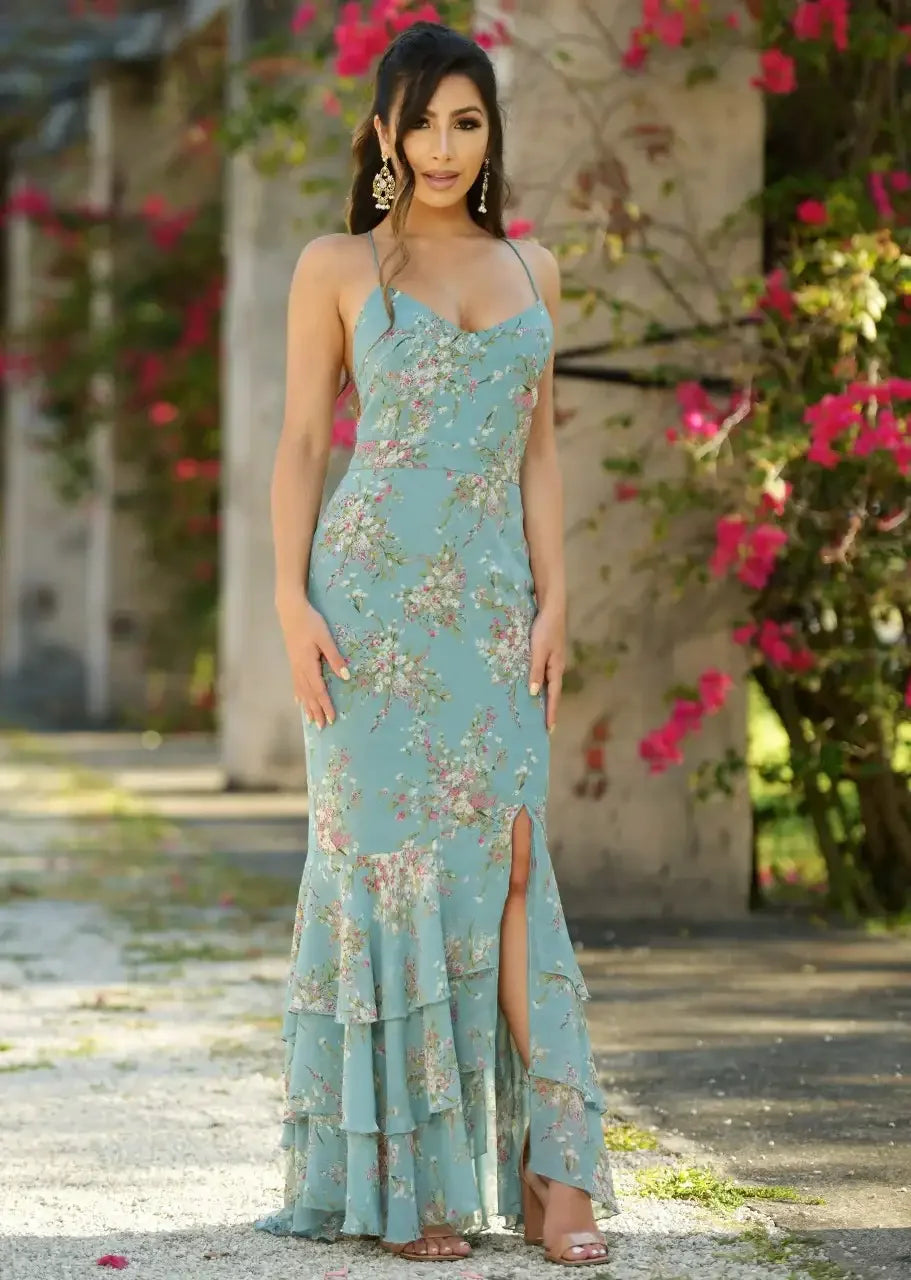 A woman in a floral, light blue gown stands elegantly, striking a confident pose. She is outdoors, surrounded by blooming pink flowers and greenery amidst weathered structures.