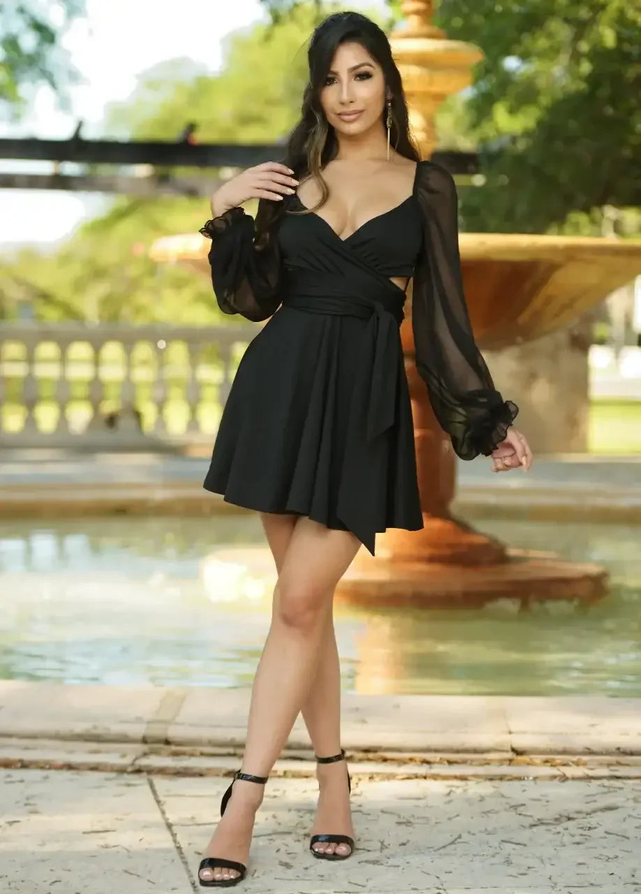 A woman stands confidently near a decorative fountain, wearing a black dress with sheer, billowy sleeves. She poses elegantly, surrounded by greenery in a bright, outdoor setting.