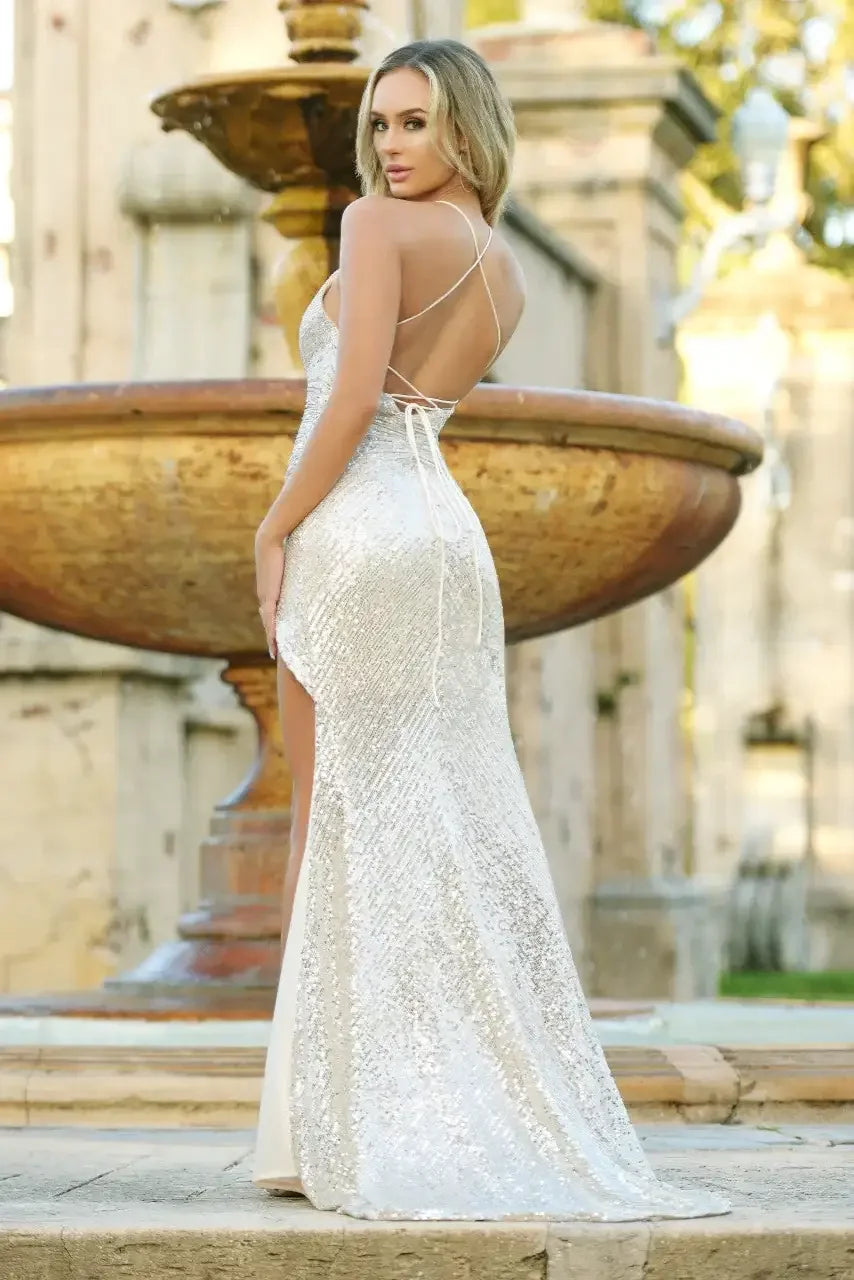 A woman in a shimmering silver gown poses gracefully near a large, decorative fountain. The gown features an open back and high slit, set against a charming outdoor backdrop.