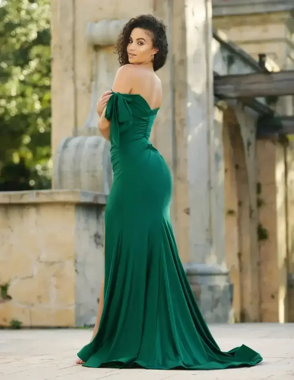 A woman in a green off-shoulder gown stands gracefully, her back turned, showcasing the dress's sleek design. The setting features soft natural lighting and stone architecture.