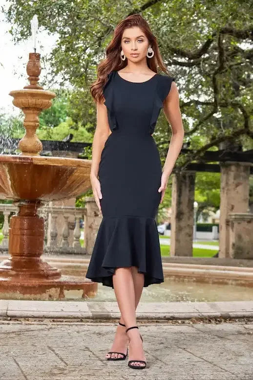 A woman stands confidently near a fountain, wearing a fitted black dress with a flared hem. She has wavy hair, large earrings, and black heels, against a backdrop of greenery.