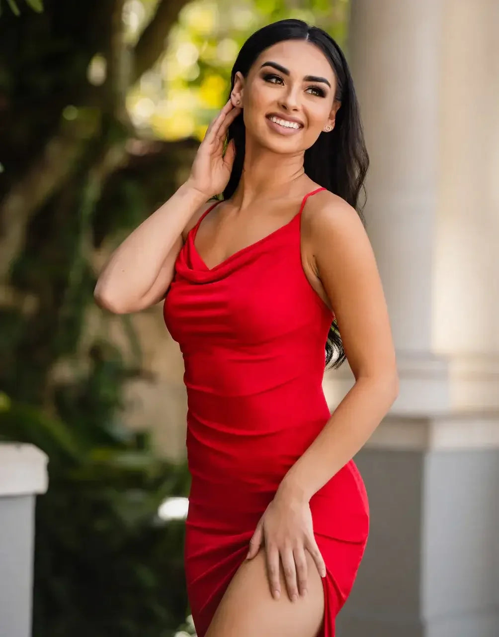 A woman in a red, form-fitting dress smiles and poses elegantly, with her hand in her hair, against a backdrop of greenery and soft architectural features, suggesting a vibrant outdoor setting.
