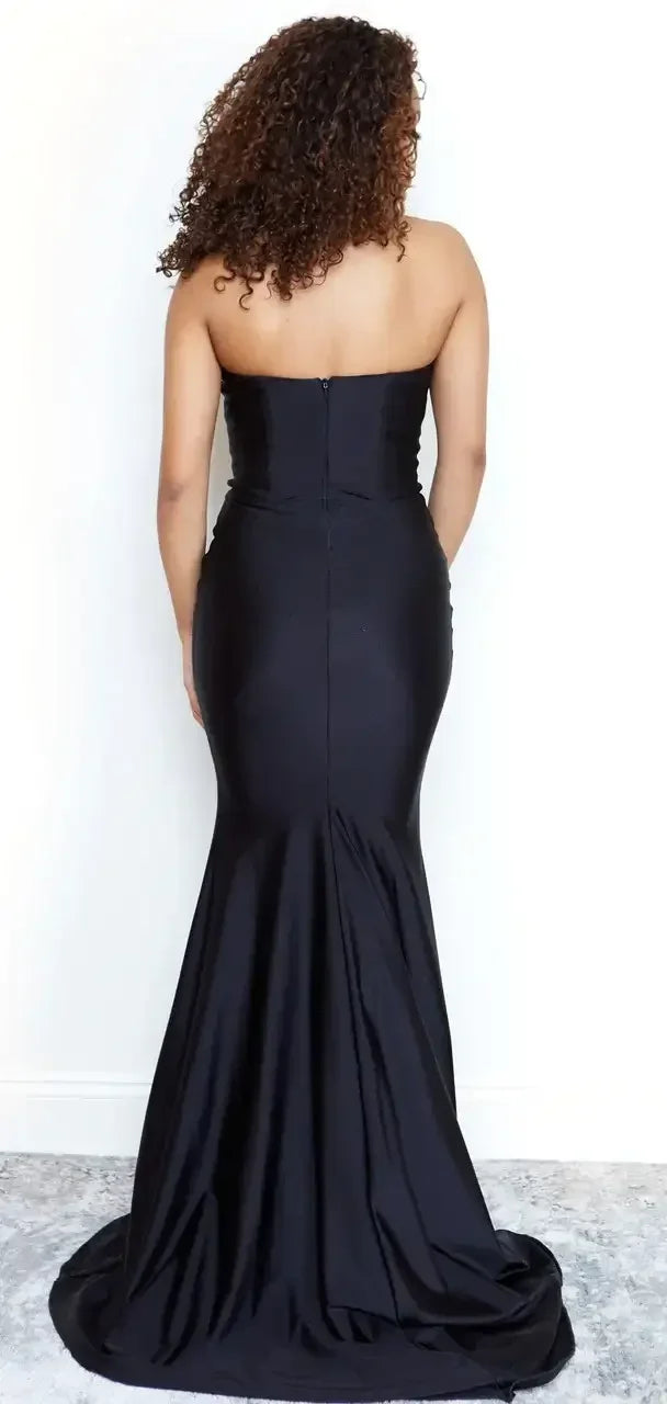 A black strapless evening gown hugs the body and flows into a flared skirt. The wearer is shown from the back, standing against a plain, light-colored wall, with a soft carpet below.