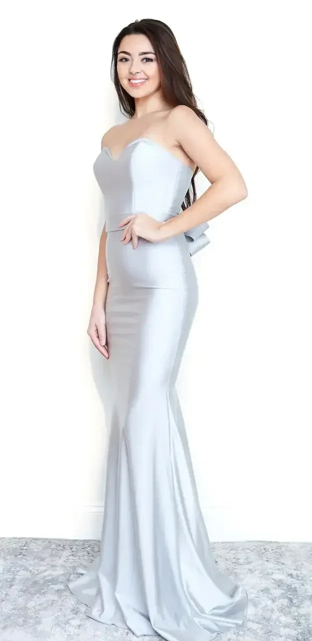 A woman stands confidently, wearing a form-fitting, light gray strapless dress with a mermaid silhouette and a bow detail on the side, against a plain white background.