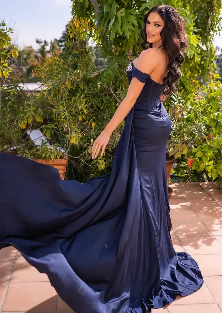 A woman in a flowing navy blue gown poses elegantly outdoors, with the dress trailing behind her. Lush greenery and potted plants create a vibrant, natural backdrop.