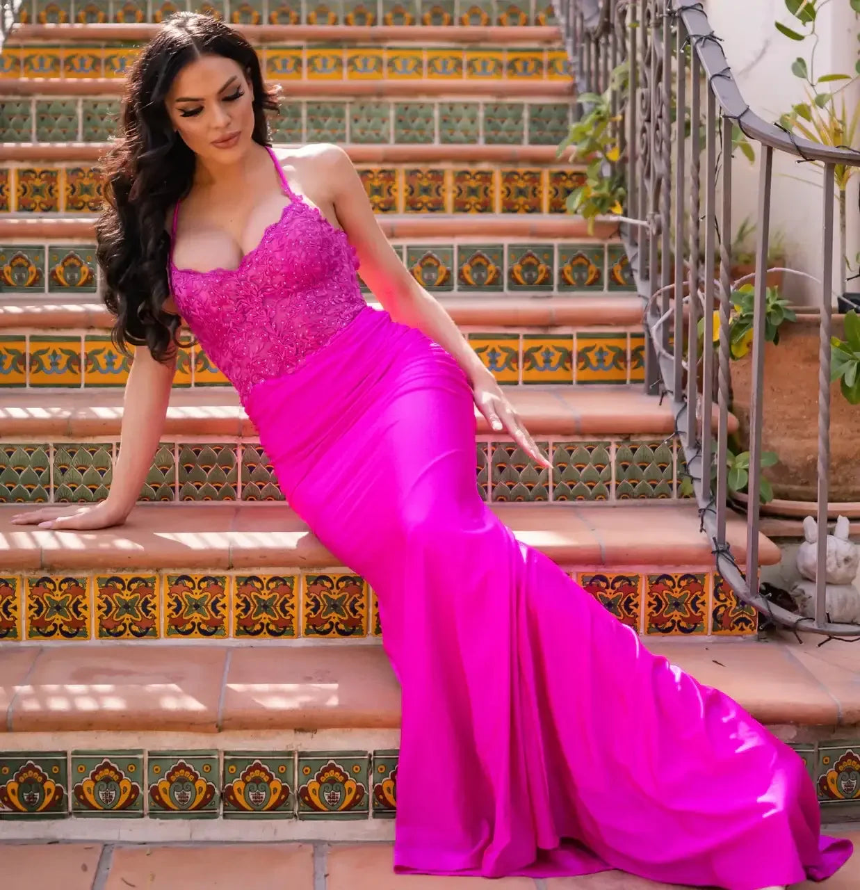 A woman in a glamorous pink dress sits gracefully on colorful tiled stairs, styled with intricate patterns, surrounded by greenery and soft sunlight, creating an elegant atmosphere.