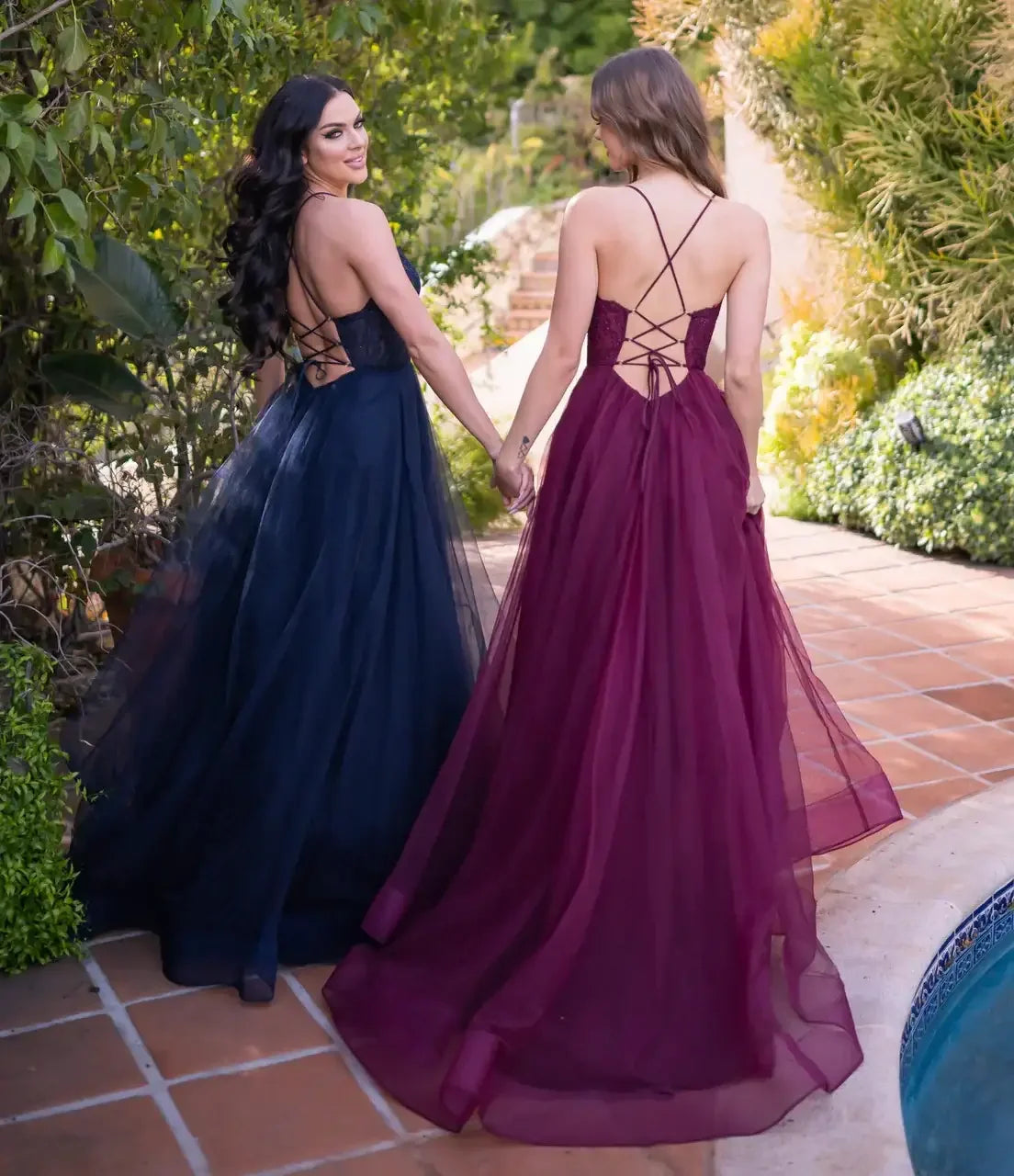 Two women in elegant evening gowns, one navy and one burgundy, are holding hands and smiling while walking together along a tiled path beside a serene pool surrounded by greenery.