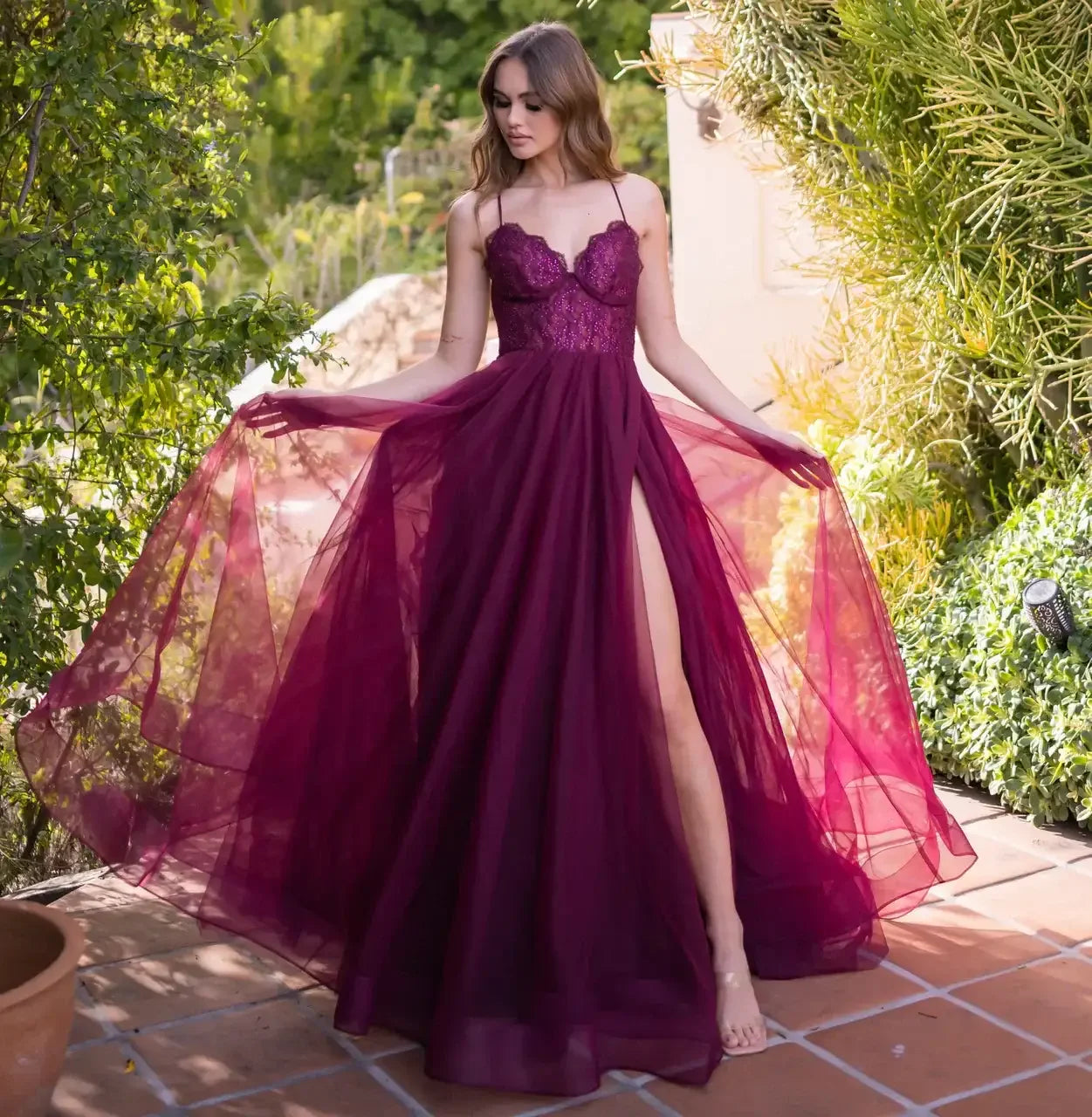 A woman in a flowing, deep burgundy gown with a thigh-high slit gracefully turns amidst a lush garden, showcasing the dress's delicate fabric against the vibrant greenery.