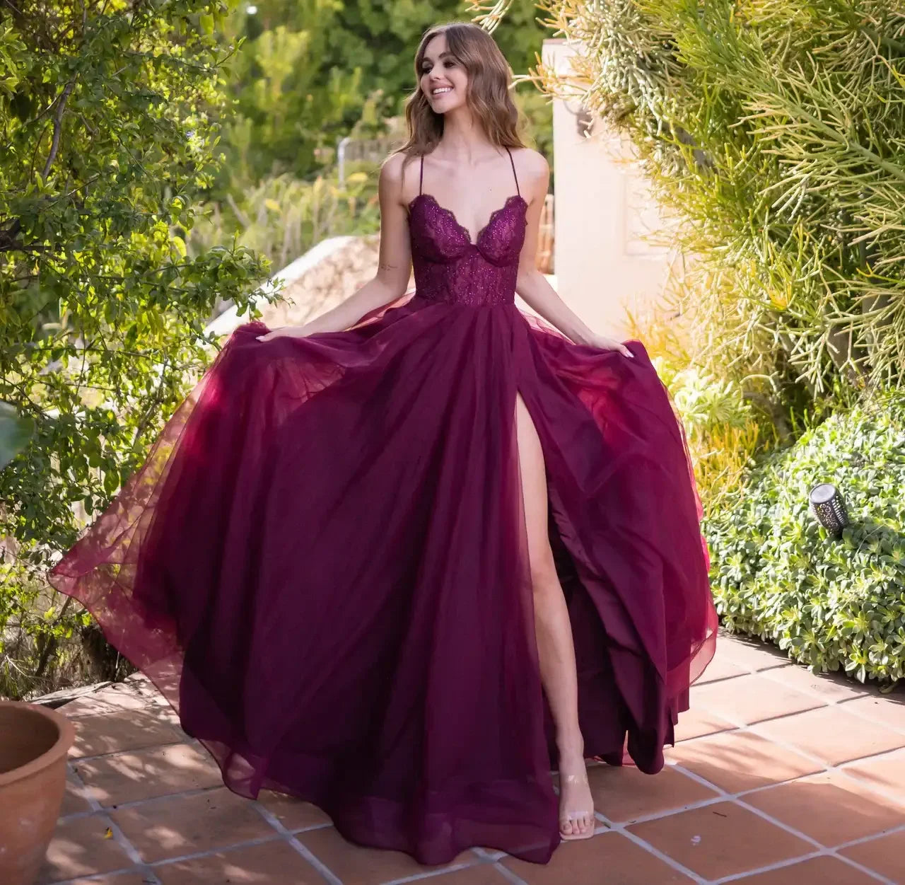 A woman in a flowing burgundy gown with a high slit twirls joyfully among greenery and stone tiles, showcasing the dress's movement and design in a sunlit outdoor setting.
