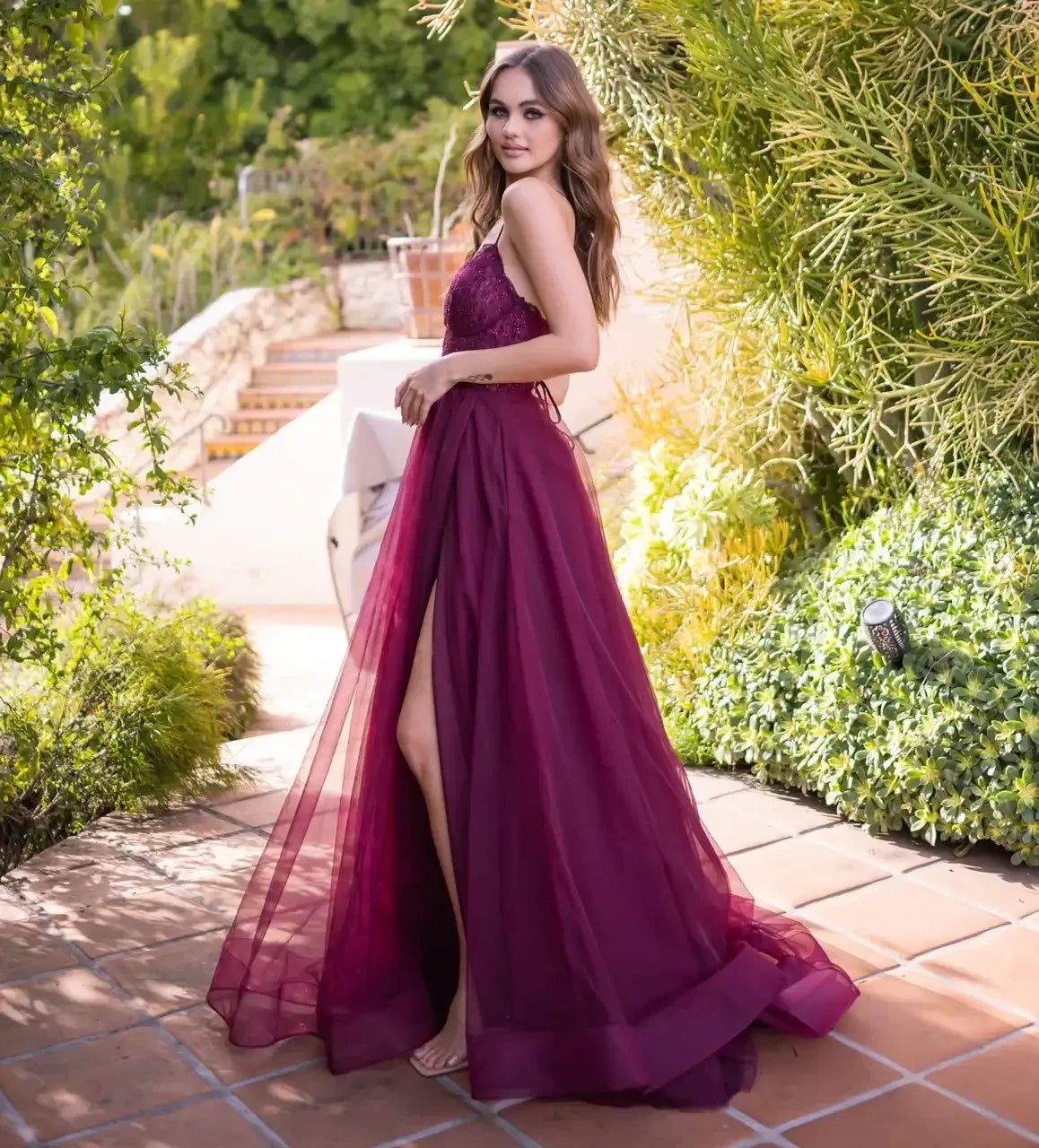 A woman in a flowing burgundy gown stands on a tiled walkway, gazing back over her shoulder. Lush greenery and soft lighting frame the serene outdoor setting.
