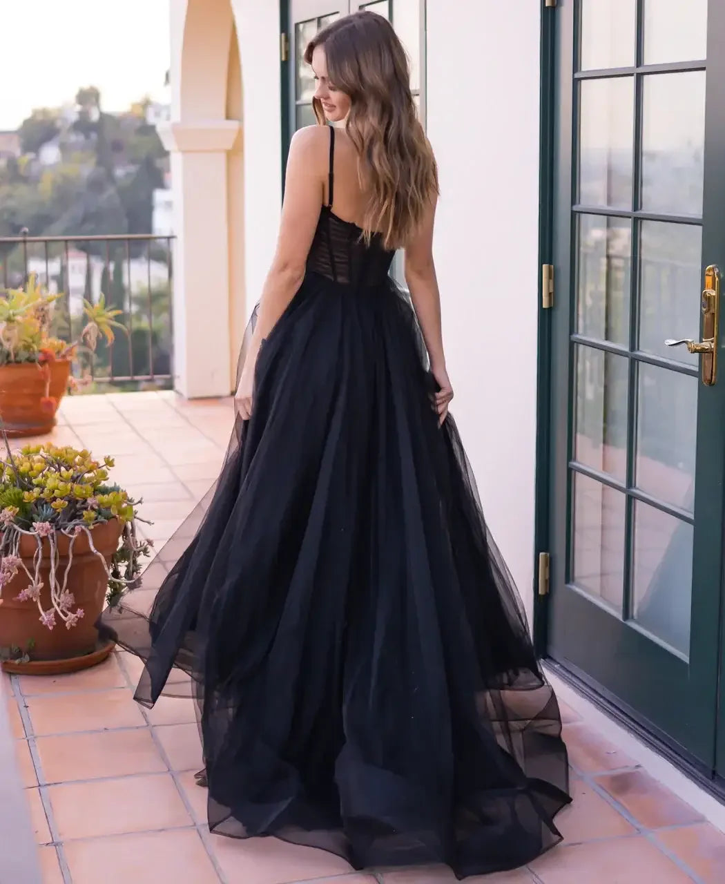 A woman in a flowing black gown stands with her back turned, gently holding the fabric. She is on a tiled patio adorned with potted plants, surrounded by a scenic outdoor view.