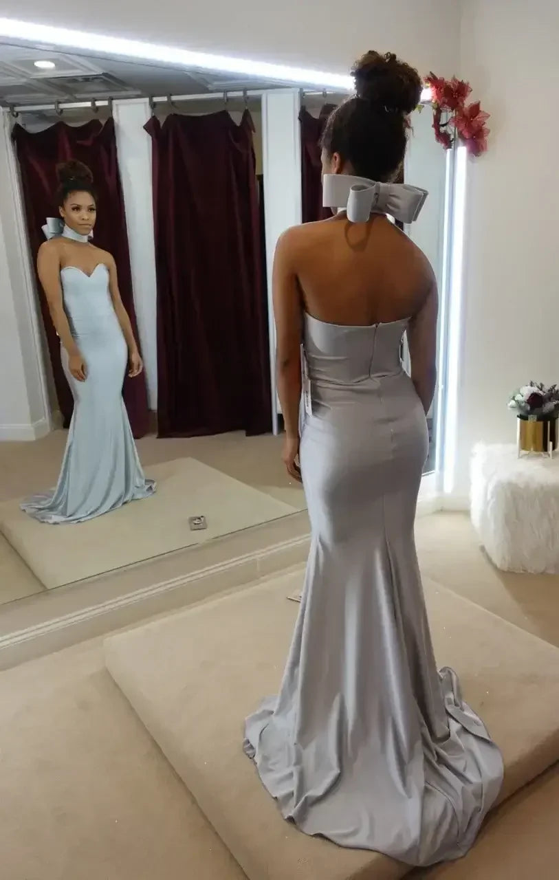 A woman admires her reflection in a mirror while wearing a sleek, silver evening gown that features a bow at the neckline, surrounded by a fitting room with burgundy curtains.