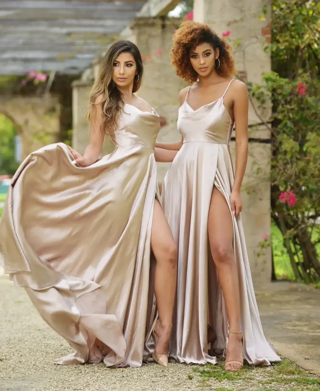 Two women stand side by side, wearing elegant, flowing satin dresses with high slits. They pose confidently in a lush garden setting, surrounded by greenery and blooming flowers.