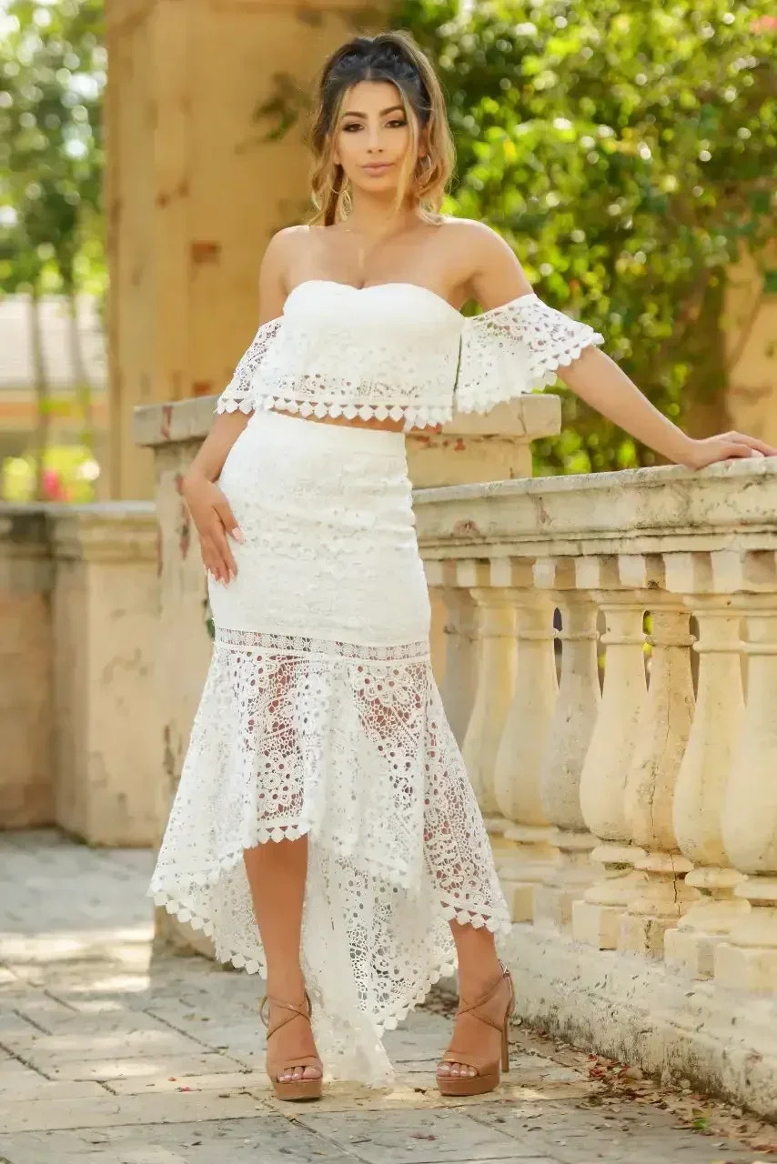 A woman in a white lace two-piece outfit stands against a stone railing, posing confidently in a lush, green outdoor setting with soft natural light enhancing the scene.