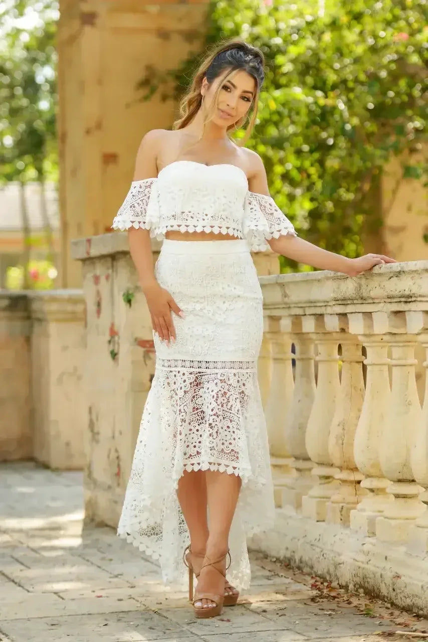 A woman in a white, lace two-piece outfit leans against a stone railing, smiling softly. The lush, green background and warm sunlight create a serene, outdoor setting.