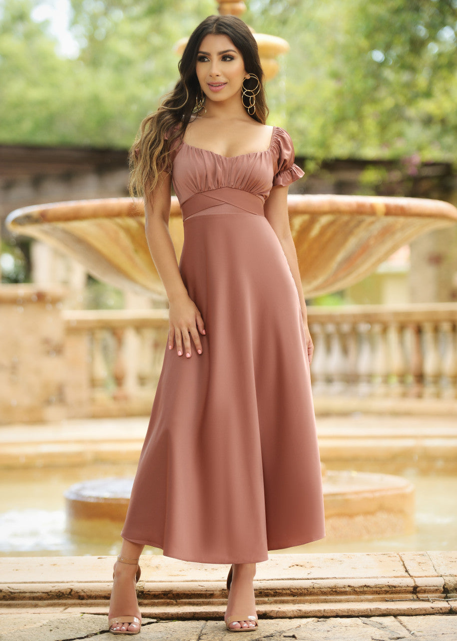 A woman stands confidently in a flowing, blush-colored dress near a fountain. Long hair cascades down, complemented by large earrings, amidst a lush, green garden setting.