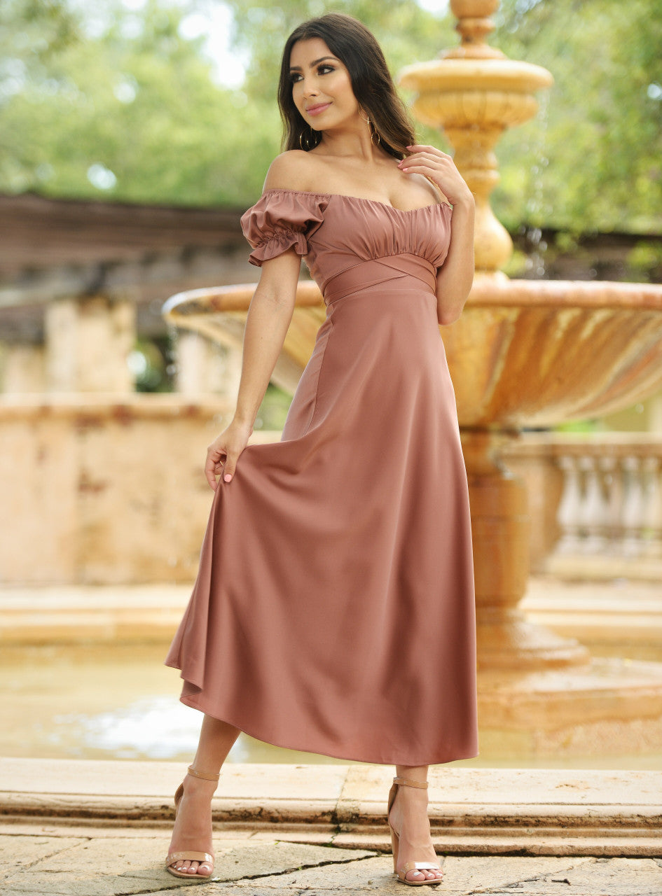 A woman in a flowing, off-the-shoulder, brown dress stands gracefully beside a stone fountain, gently holding the fabric while smiling, surrounded by lush greenery and soft sunlight.